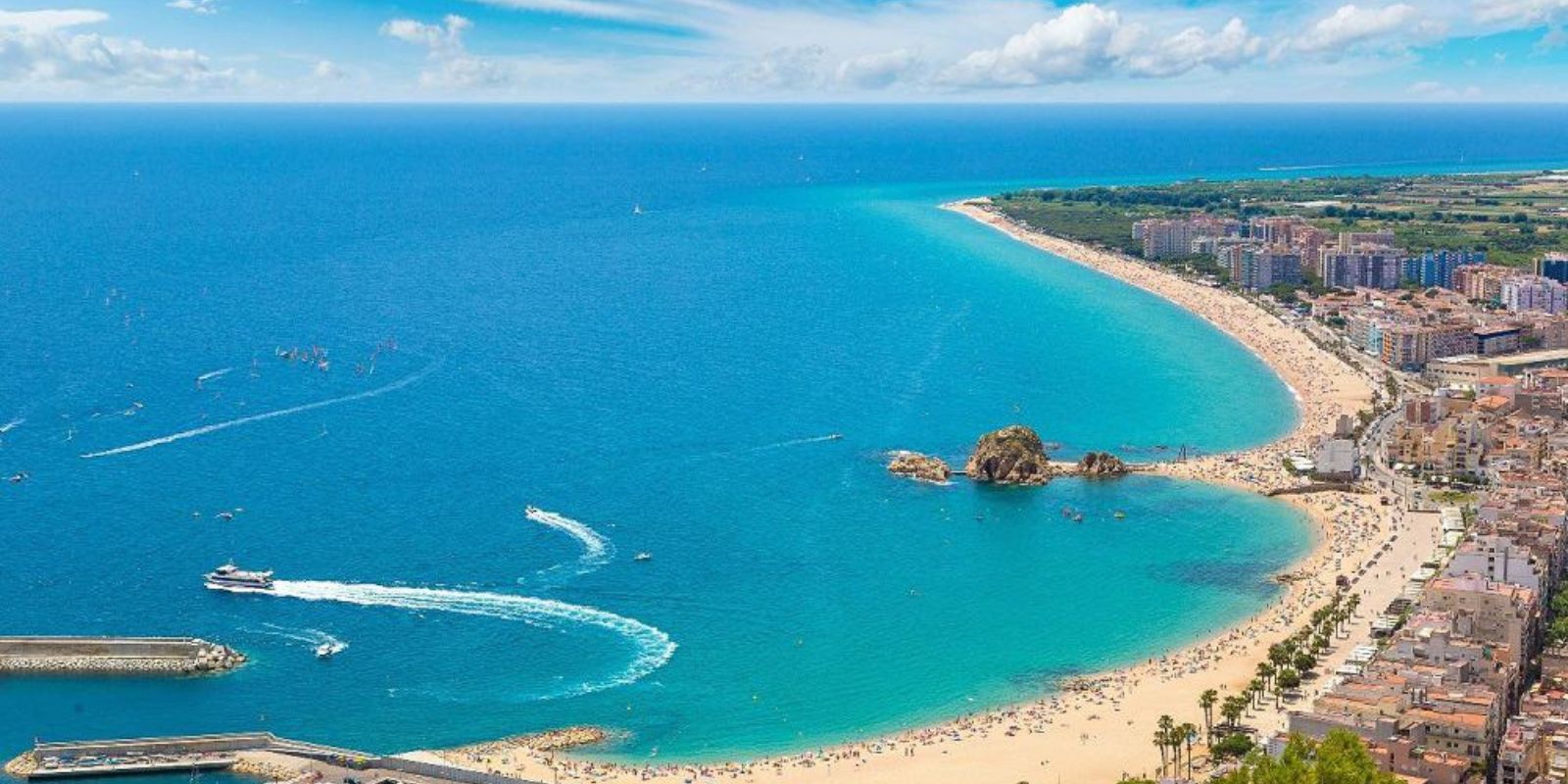 Belle plage dorée bordée par la mer turquoise et une promenade animée, avec bateaux au port et bâtiments côtiers sous un ciel bleu. intégrer le mot-clé comment organiser séjour espagne de façon naturelle.