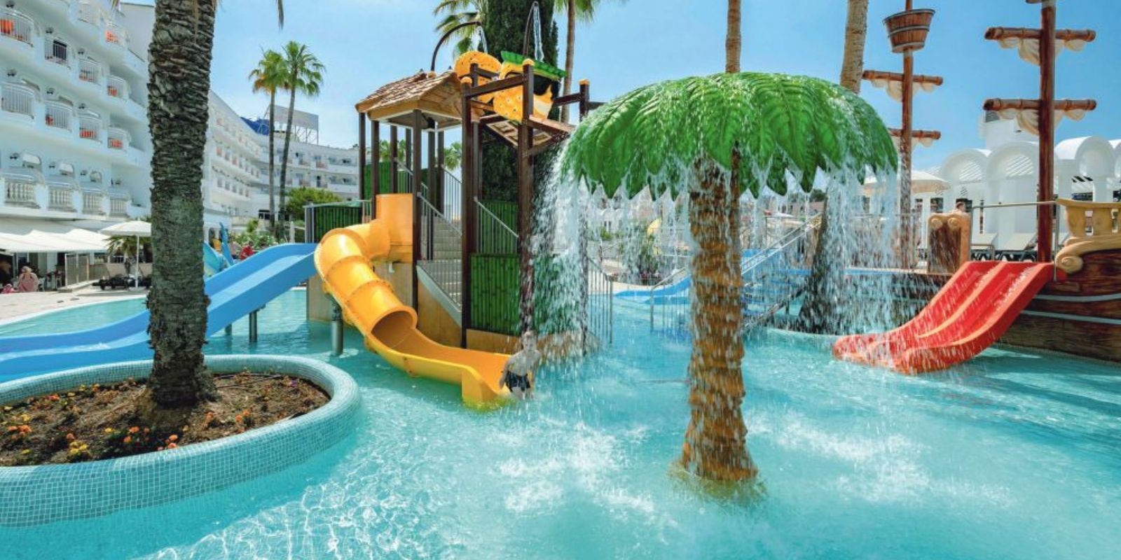 Pic d’un toboggan rouge dans une pataugeoire bleue, hôtel all inclusive Espagne en arrière-plan avec transats et bâtiment en arcades.